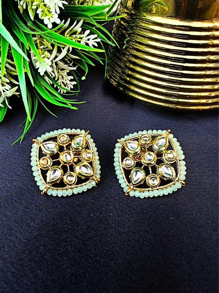 Sharvisha Mint Green Earrings: Square-shaped Kundan earrings with teardrop white Kundan accents, surrounded by Mint Green crystal stones, displayed on a Black surface with flowers & Leaves in the background.