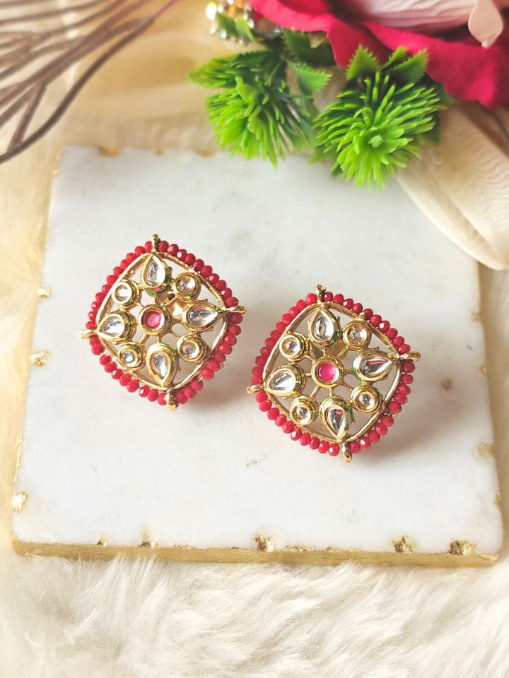Sharvisha Red Earrings: Square-shaped Kundan earrings with teardrop white Kundan accents, surrounded by red crystal stones, displayed on a marble-like surface with flowers in the background.