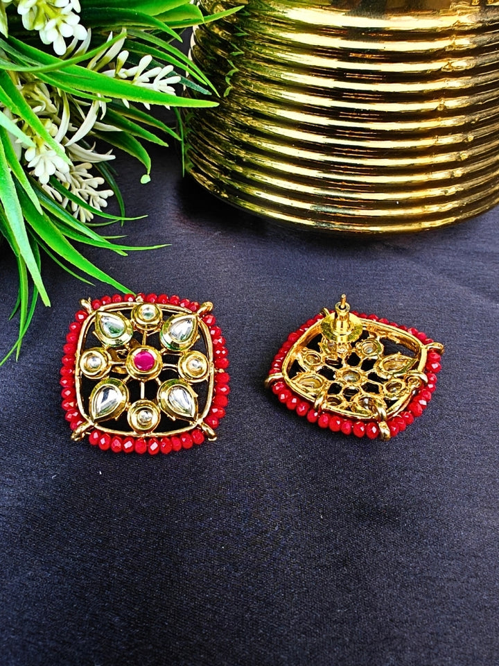 Sharvisha Red Earrings: Square-shaped Kundan earrings with teardrop white Kundan accents, surrounded by red crystal stones, displayed on a Black surface with flowers & Leaves With Accent in the background.