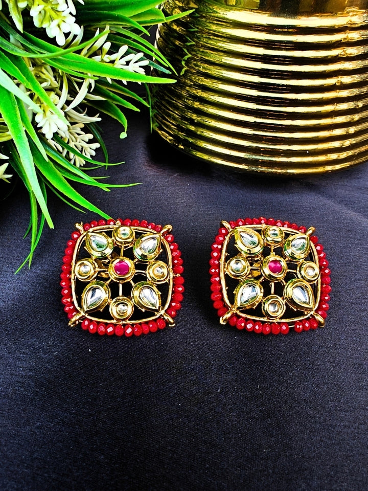 Sharvisha Red Earrings: Square-shaped Kundan earrings with teardrop white Kundan accents, surrounded by red crystal stones, displayed on a Black surface with flowers & Leaves With Accent in the background.