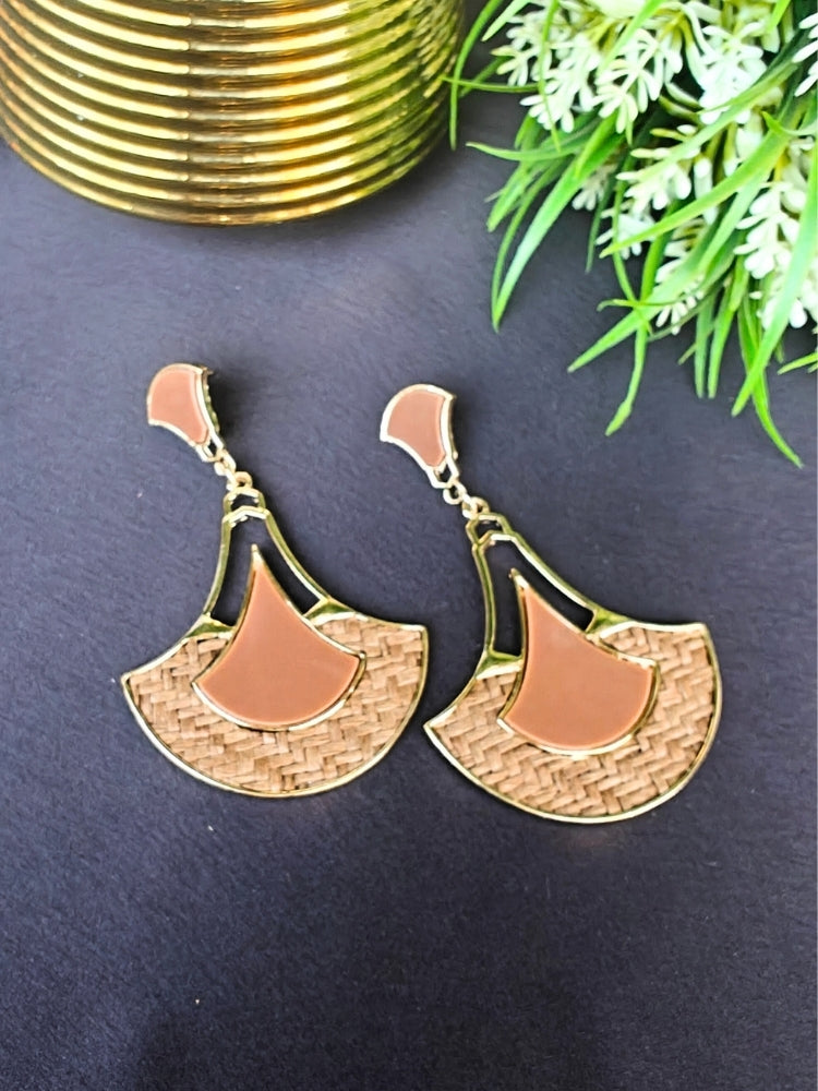 The Shefali Brown Earrings feature a fan shape long earrings with gold edges, tan and light brown geometric accents, and woven textured centers—an elegant ethnic accessory shown on a dark surface near a gold-striped vase and leafy plant.