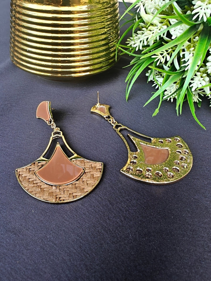 The Shefali Brown Earrings feature a fan shape long earrings with gold edges, tan and light brown geometric accents, and woven textured centers—an elegant ethnic accessory shown on a dark surface near a gold-striped vase and leafy plant.
