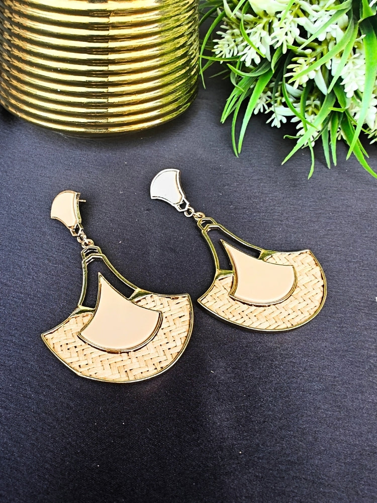 The Shefali Cream Earrings feature a fan shape long earrings with gold edges, tan and light Cream geometric accents, and woven textured centers—an elegant ethnic accessory shown on a dark surface near a gold-striped vase and leafy plant.