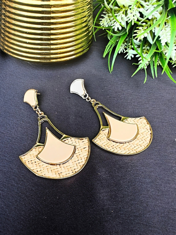The Shefali Cream Earrings feature a fan shape long earrings with gold edges, tan and light Cream geometric accents, and woven textured centers—an elegant ethnic accessory shown on a dark surface near a gold-striped vase and leafy plant.