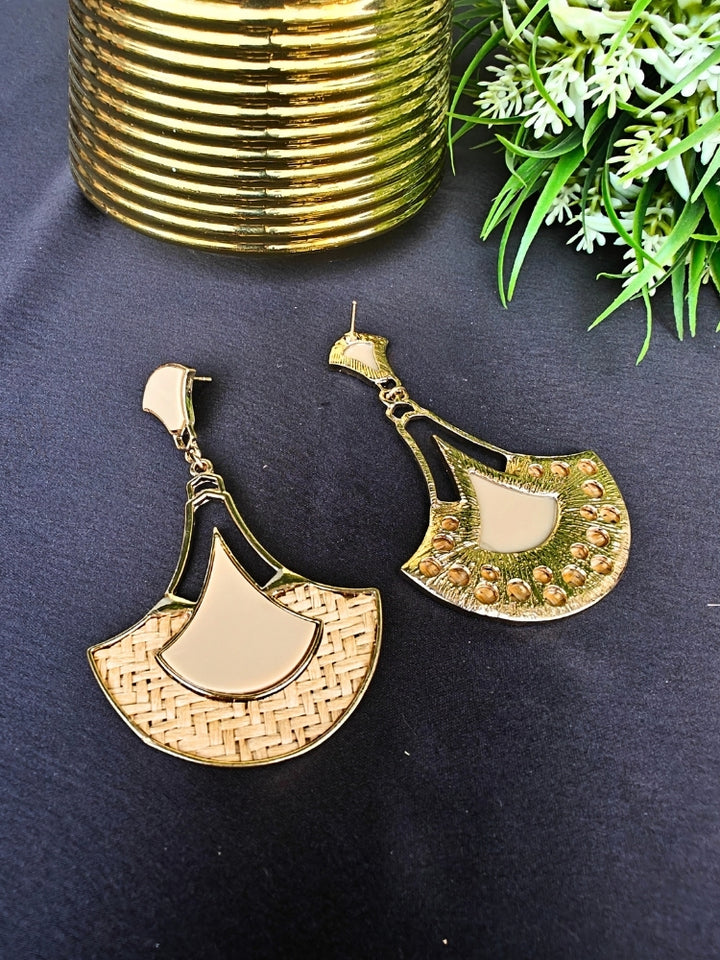 The Shefali Cream Earrings feature a fan shape long earrings with gold edges, tan and light Cream geometric accents, and woven textured centers—an elegant ethnic accessory shown on a dark surface near a gold-striped vase and leafy plant.
