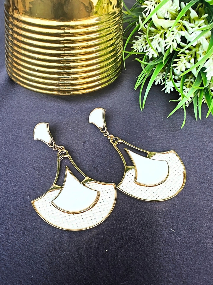 The Shefali White Earrings feature a fan shape long earrings with gold edges, tan and White geometric accents, and woven textured centers—an elegant ethnic accessory shown on a dark surface near a gold-striped vase and leafy plant.