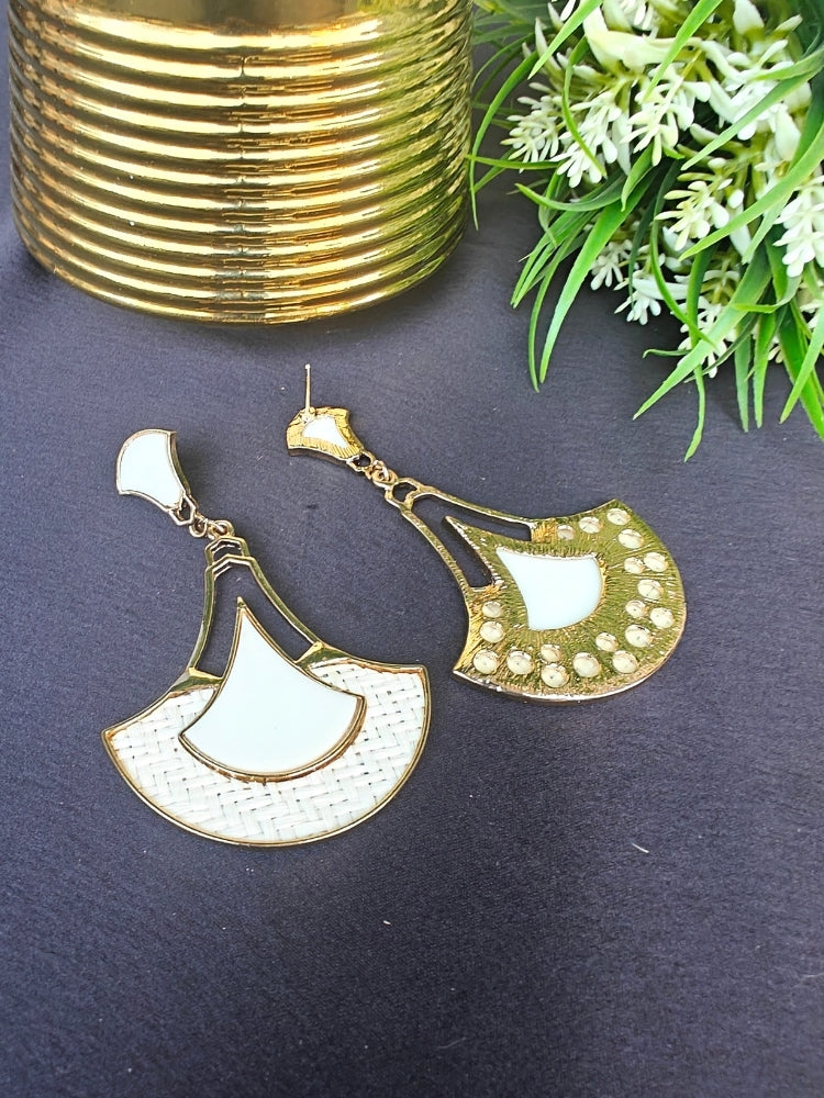 The Shefali White Earrings feature a fan shape long earrings with gold edges, tan and White geometric accents, and woven textured centers—an elegant ethnic accessory shown on a dark surface near a gold-striped vase and leafy plant.