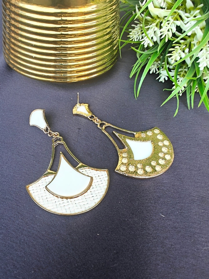 The Shefali White Earrings feature a fan shape long earrings with gold edges, tan and White geometric accents, and woven textured centers—an elegant ethnic accessory shown on a dark surface near a gold-striped vase and leafy plant.