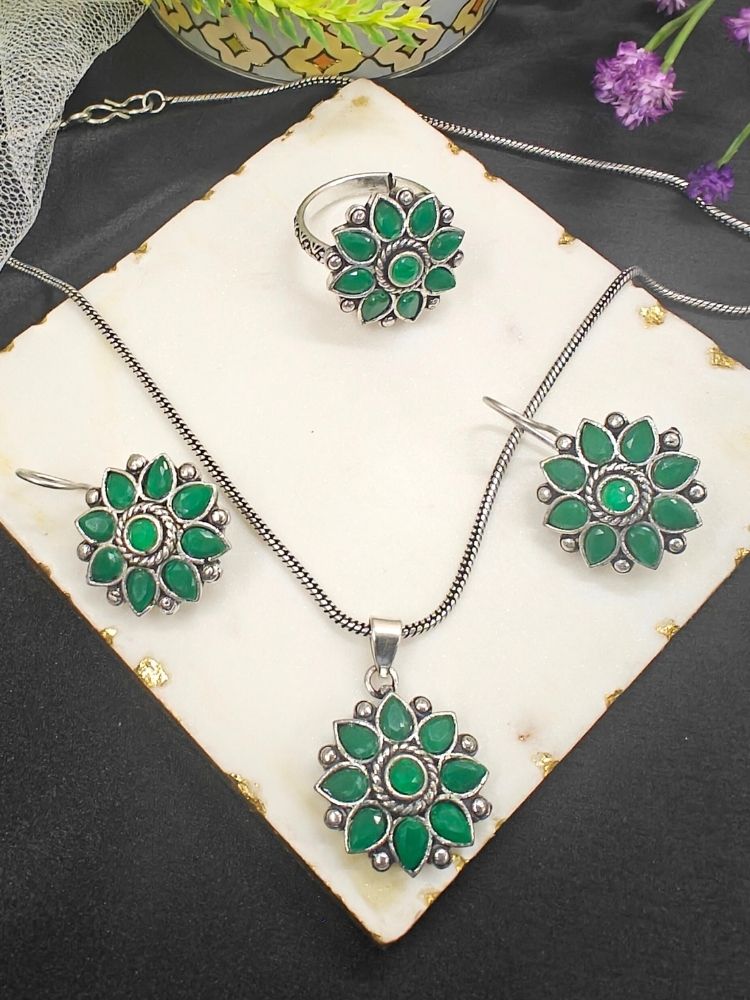 The Shivangi Green Pendant Set with Ring features a oxidized silver ethnic floral design with green gemstones, including a pendant necklace, ring, and earrings—ideal for wedding ceremonies. Shown on a white tile with gold edges and purple flowers.