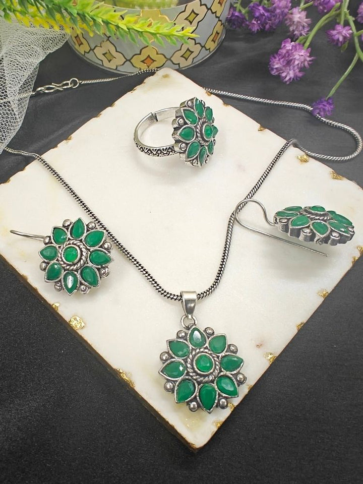 The Shivangi Green Pendant Set with Ring features a oxidized silver ethnic floral design with green gemstones, including a pendant necklace, ring, and earrings—ideal for wedding ceremonies. Shown on a white tile with gold edges and purple flowers.