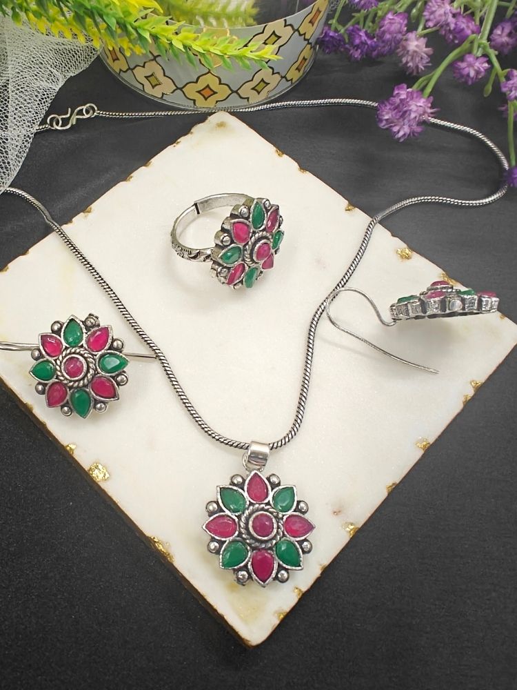 The Shivangi Magenta & Pink Pendant Set with Ring features a oxidized silver ethnic floral design with Magenta & Pink gemstones, including a pendant necklace, ring, and earrings—ideal for wedding ceremonies. Shown on a white tile with gold edges and purple flowers