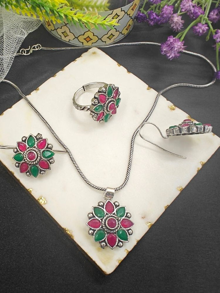The Shivangi Magenta & Pink Pendant Set with Ring features a oxidized silver ethnic floral design with Magenta & Pink gemstones, including a pendant necklace, ring, and earrings—ideal for wedding ceremonies. Shown on a white tile with gold edges and purple flowers