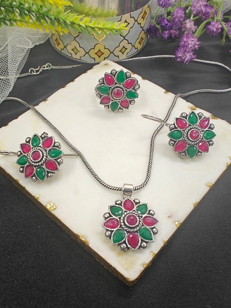 The Shivangi Magenta & Pink Pendant Set with Ring features a oxidized silver ethnic floral design with Magenta & Pink gemstones, including a pendant necklace, ring, and earrings—ideal for wedding ceremonies. Shown on a white tile with gold edges and purple flowers