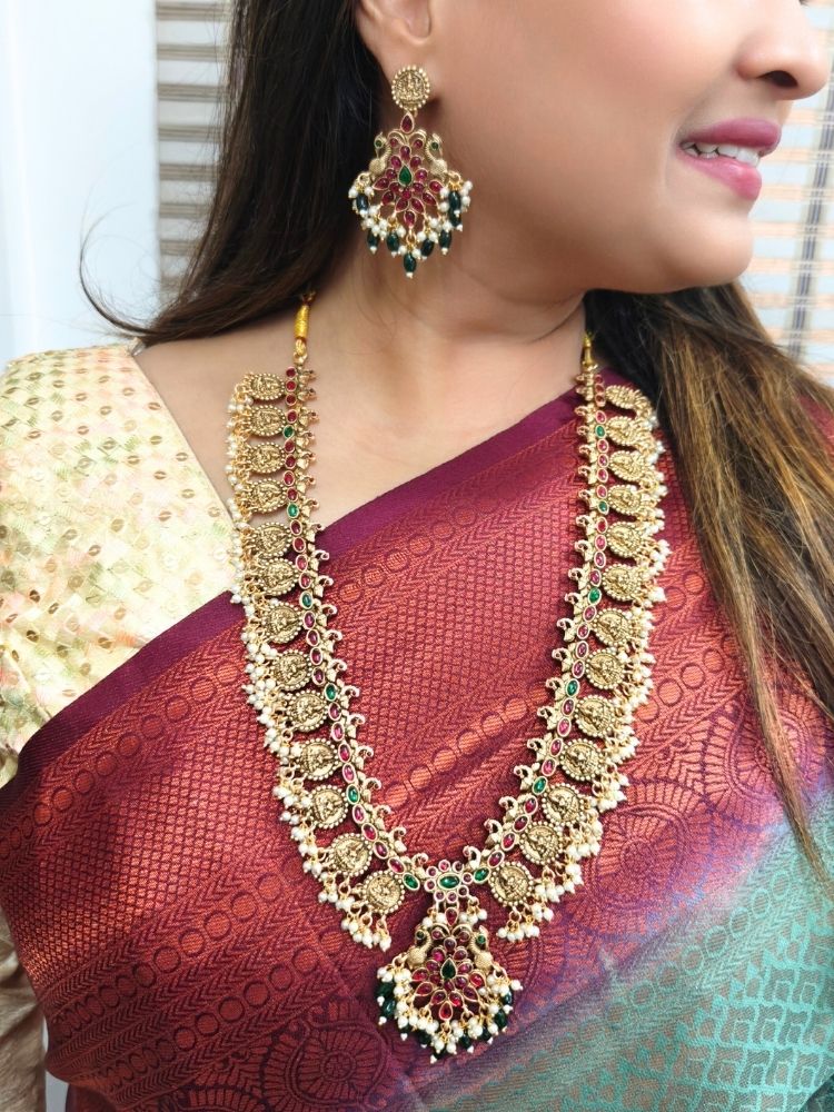 A woman in a red and gold sari with a beige blouse smiles, adorned with the Shraddha Temple Necklace Set featuring Maa Laxmi motifs all around the necklace and intricate details highlighted by red and green gemstones.