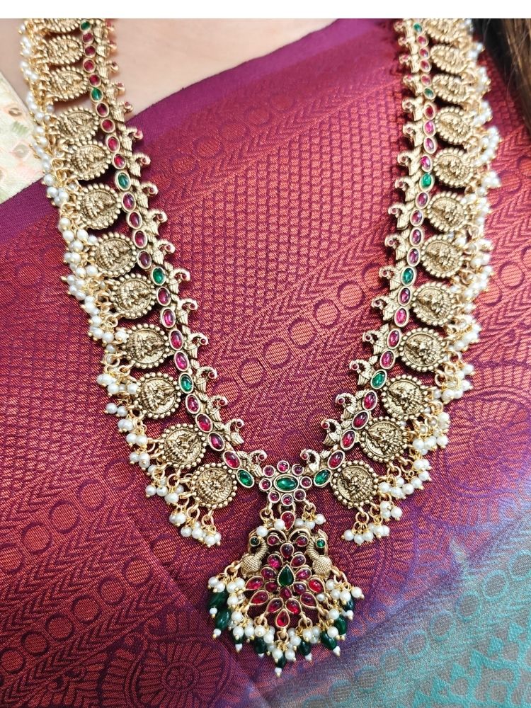 A woman in a red and gold sari with a beige blouse smiles, adorned with the Shraddha Temple Necklace Set featuring Maa Laxmi motifs all around the necklace and intricate details highlighted by red and green gemstones.