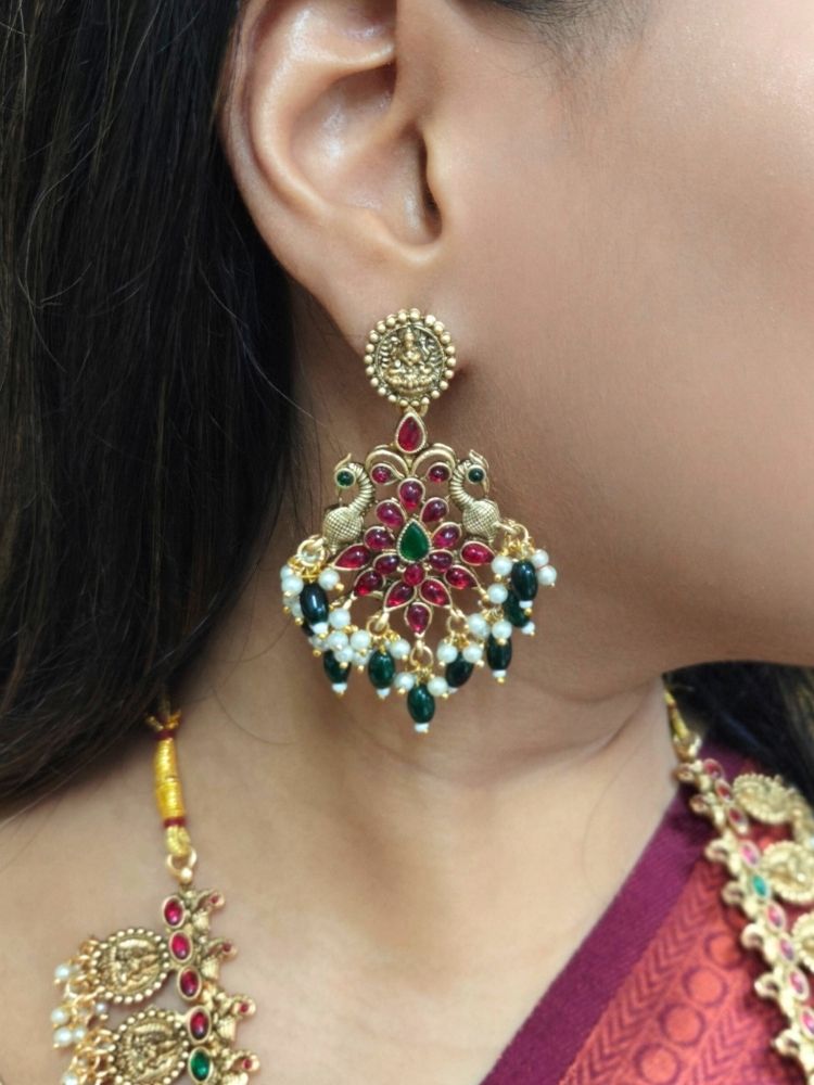 A woman in a red and gold sari with a beige blouse smiles, adorned with the Shraddha Temple Necklace Set featuring Maa Laxmi motifs all around the necklace and intricate details highlighted by red and green gemstones.