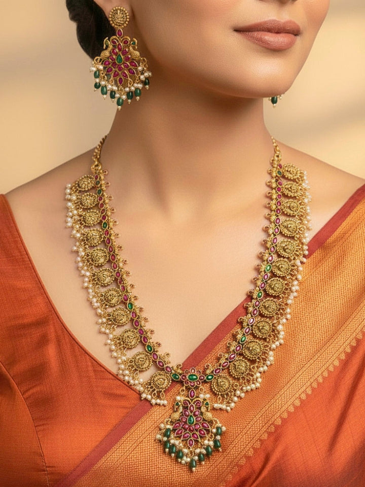 A woman wearing the Shraddha Temple Necklace Set featuring a traditional gold Long necklace and matching earrings with   Pearls, Colored Stones and Mehendi Polish . Bride wearing a handcrafted temple artificial jewelry set for reception night. Close-up of detailed temple carvings in artificial wedding jewelry.