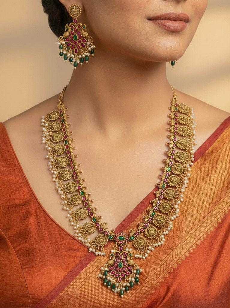A woman wearing the Shraddha Temple Necklace Set featuring a traditional gold Long necklace and matching earrings with   Pearls, Colored Stones and Mehendi Polish . Bride wearing a handcrafted temple artificial jewelry set for reception night. Close-up of detailed temple carvings in artificial wedding jewelry.