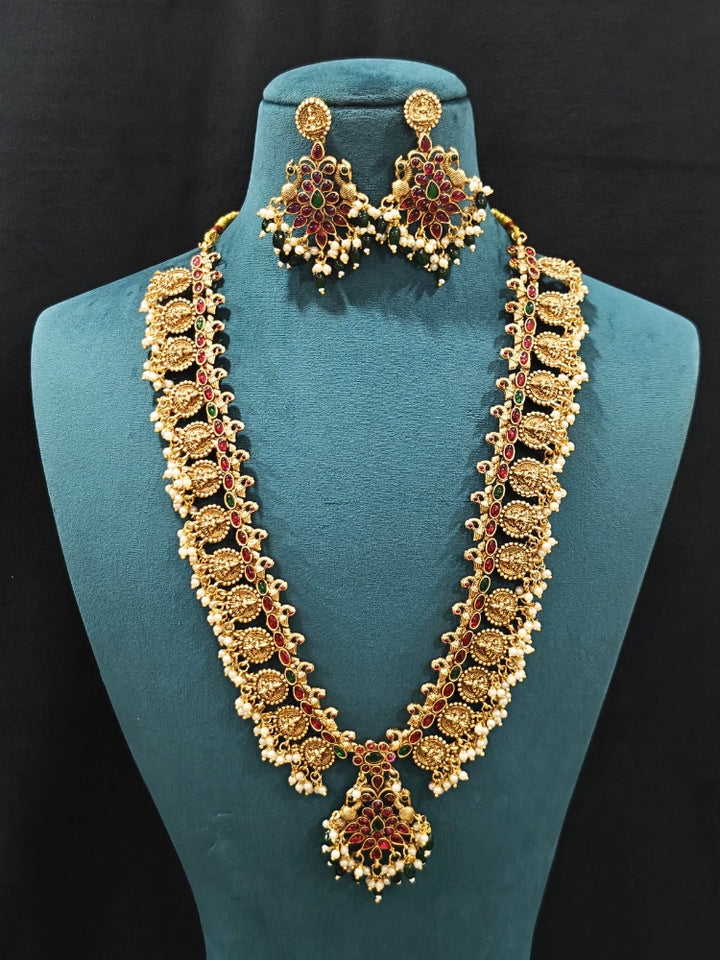 The Shraddha Temple Necklace Set featuring a traditional gold Long necklace and matching earrings with   Pearls, Colored Stones and Mehendi Polish .  Close-up of detailed temple carvings in artificial wedding jewelry. Festive Diwali look completed with a traditional temple necklace set.