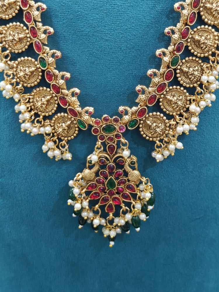 The Shraddha Temple Necklace Set featuring a traditional gold Long necklace and matching earrings with   Pearls, Colored Stones and Mehendi Polish .  Close-up of detailed temple carvings in artificial wedding jewelry. Festive Diwali look completed with a traditional temple necklace set.