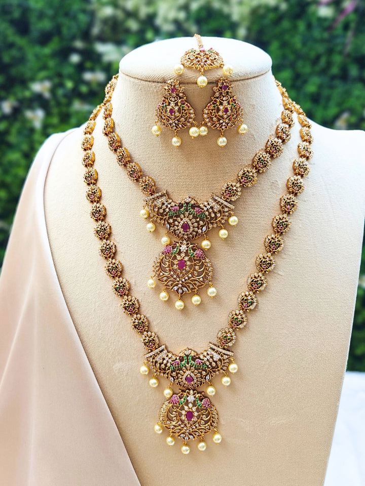 The Shweta Necklace Set is displayed on a mannequin, featuring ornate two-layered gold necklaces with red and green stones, American Diamonds, pearl drops, and matching earrings, set against a blurred greenery background.