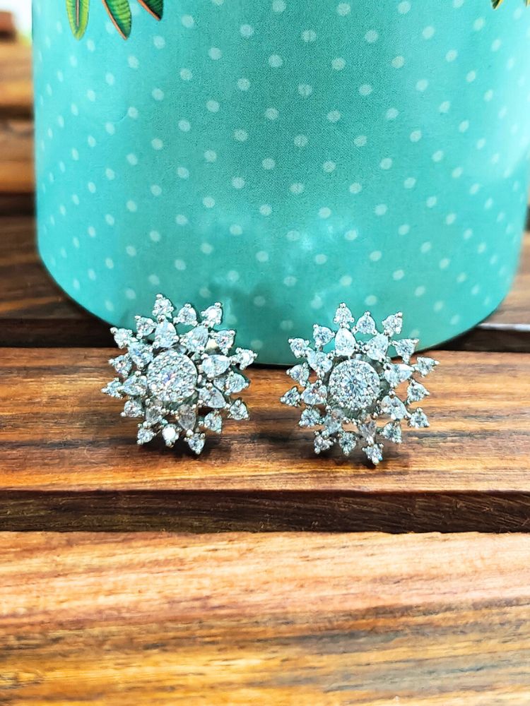 The Silver Dahlia stud Earrings are chic, flower-shaped Silver stud earrings adorned with American diamonds, elegantly displayed on a wooden surface against a green polka-dotted background.