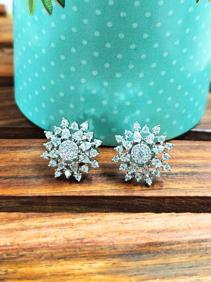 The Silver Dahlia stud Earrings are chic, flower-shaped Silver stud earrings adorned with American diamonds, elegantly displayed on a wooden surface against a green polka-dotted background.