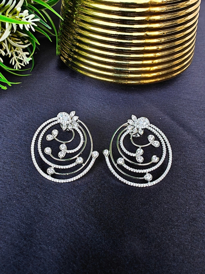 The Silver Maria Earrings feature sparkling circular American Diamonds with intricate spiral designs and clear stones, beautifully displayed on a black surface beside gold decor and green foliage. American Diamond Earrings