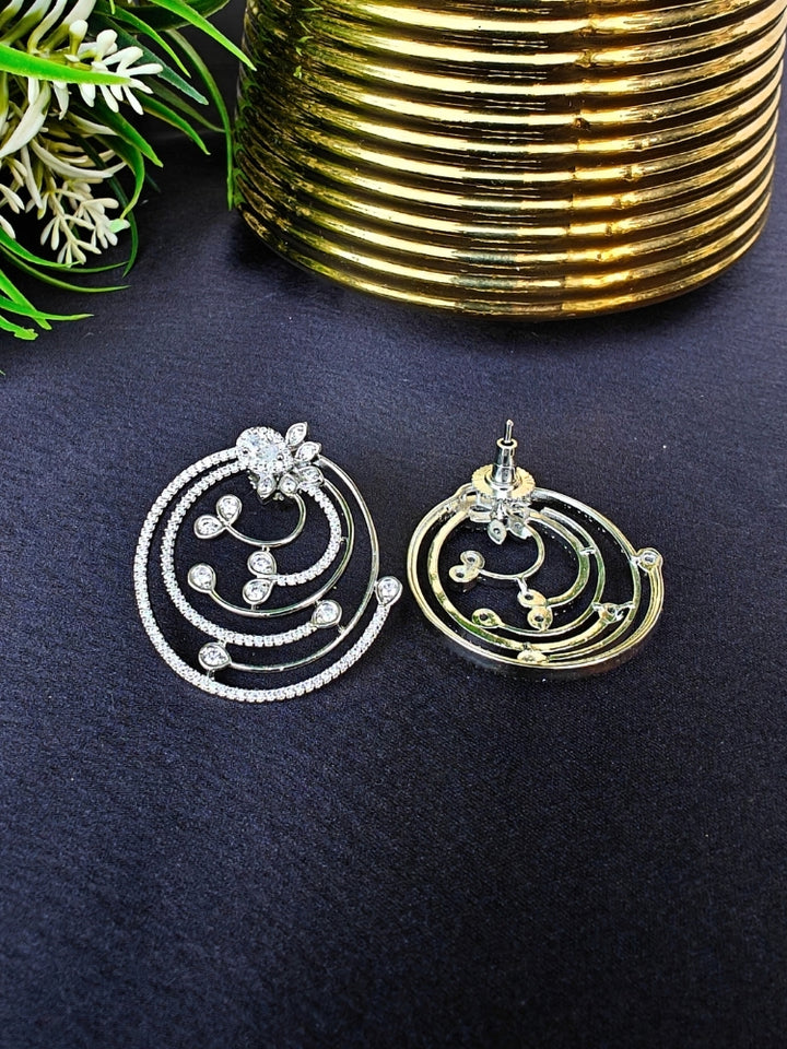 The Silver Maria Earrings feature sparkling circular American Diamonds with intricate spiral designs and clear stones, beautifully displayed on a black surface beside gold decor and green foliage. American Diamond Earrings