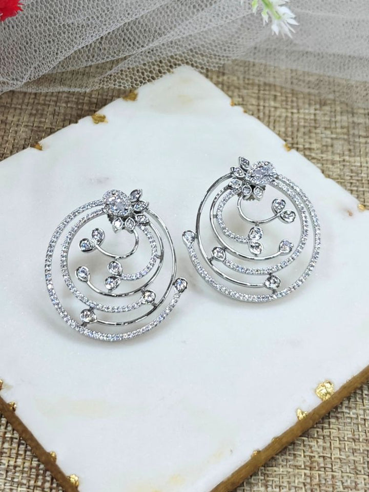 The Silver Maria Earrings feature sparkling circular American Diamonds with intricate spiral designs and clear stones, beautifully displayed on a White surface beside Flower decor and white  Net. American Diamond Earrings
