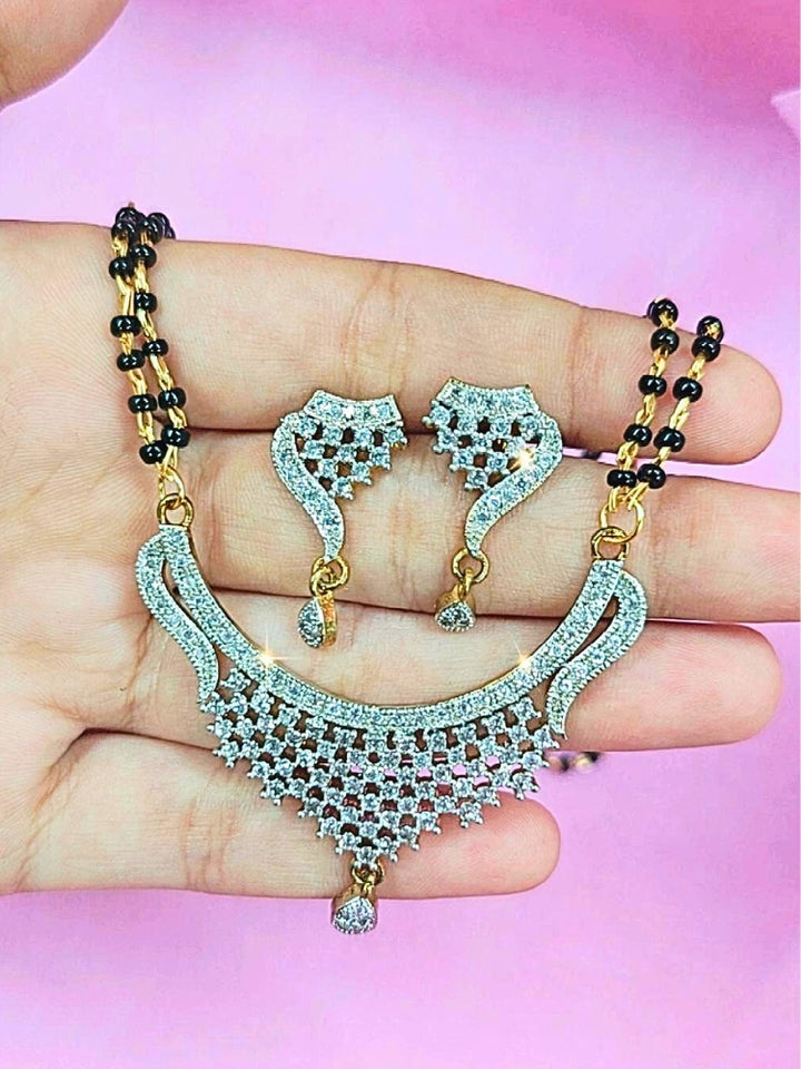 A hand holds the Simmi Mangalsutra—a gold necklace with black mangalsutra beads and matching earrings, pendant featuring sparkling American Diamonds—set against a soft pink background, perfect for your wedding ceremony dress. Diamond mangalsutra. Modern mangalsutra. 