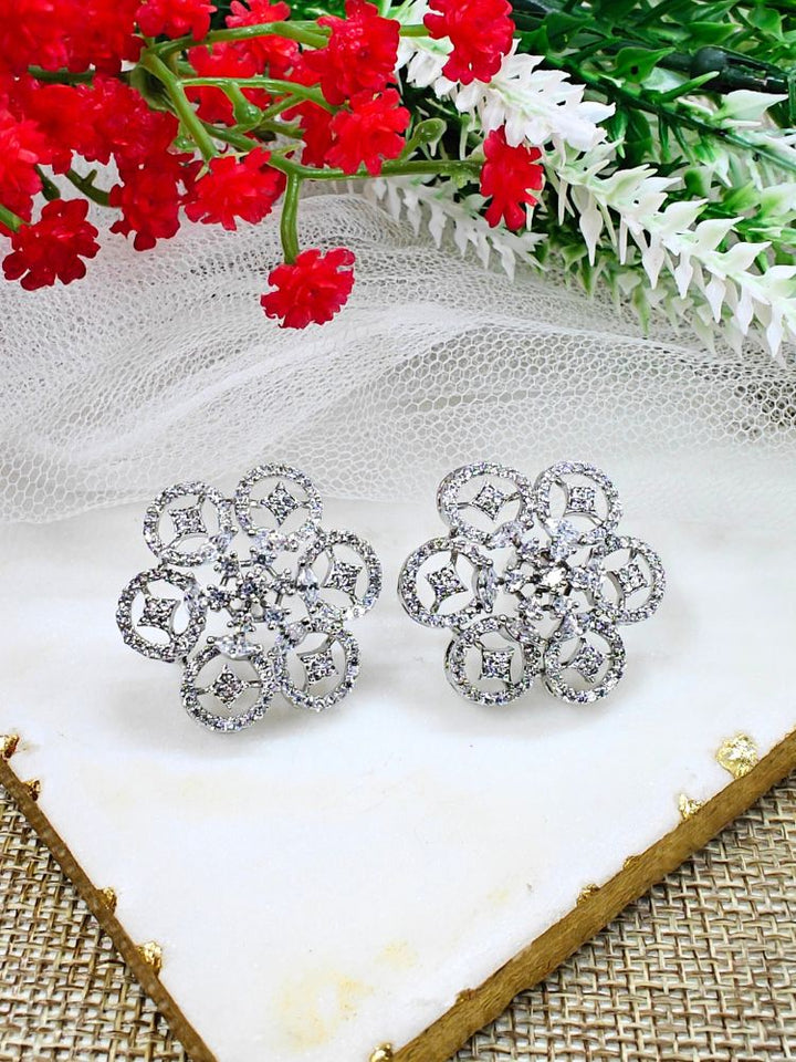 The Sloan Earrings are sparkling silver earrings with American Diamonds and intricate floral designs, showcased on a white fabric backdrop alongside red and white artificial flowers and greenery.