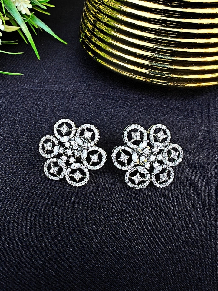The Sloan Earrings are sparkling silver earrings with American Diamonds and intricate floral designs, showcased on a white fabric backdrop alongside red and white artificial flowers and Golden accent.