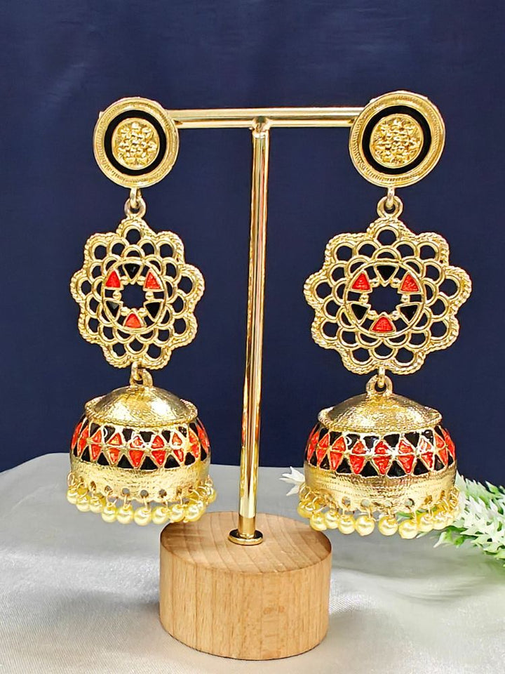 The Suhani Earrings are ornate gold and red jhumka earrings featuring intricate filigree, dome-shaped drops, and small gold beads, elegantly presented on a gold stand with a wooden base against a dark background.