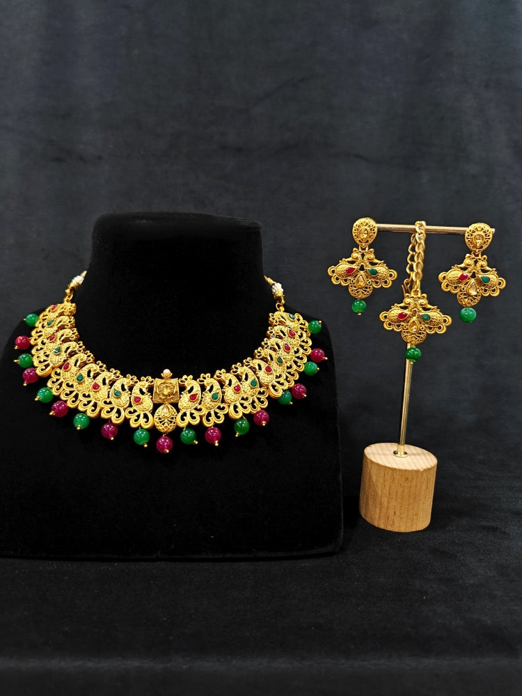 Close-up of an ornate traditional (Gold Polish) Collar jewellery set and earrings and maangtikka with Red & Green Colored Stone and Beads. Festive party wear look featuring a statement artificial Colored Stone jewelry piece. Woman wearing lightweight artificial Colored Stone jewelry for long wedding functions.