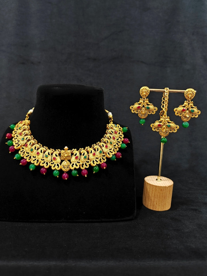 Close-up of an ornate traditional (Gold Polish) Collar jewellery set and earrings and maangtikka with Red & Green Colored Stone and Beads. Festive party wear look featuring a statement artificial Colored Stone jewelry piece. Woman wearing lightweight artificial Colored Stone jewelry for long wedding functions.