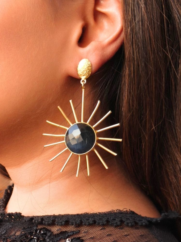 Close-up of a womans ear wearing Sun Earrings—large, gold sunburst-style with a black stone center and paired with a textured gold stud—complementing her brown hair and black top, ideal for pairing with a wedding ceremony dress.