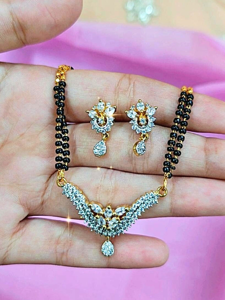 A hand holds the Supriya Mangalsutra—a gold necklace with black mangalsutra beads and matching earrings, pendant featuring sparkling American Diamonds—set against a soft pink background, perfect for your wedding ceremony dress. Diamond mangalsutra. Modern mangalsutra. 