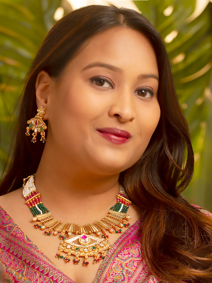 A woman smiles softly wearing the Svarlina Green & Pink Necklace Set, featuring a Kundan necklace with green and red crystal stones, matching earrings, and a gold-embroidered pink traditional outfit—ideal elegant party wear jewelry.