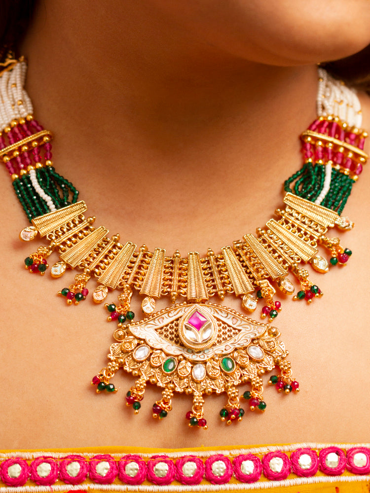 A woman smiles softly wearing the Svarlina Green & Pink Necklace Set, featuring a Kundan necklace with green and red crystal stones, matching earrings, and a gold-embroidered pink traditional outfit—ideal elegant party wear jewelry.