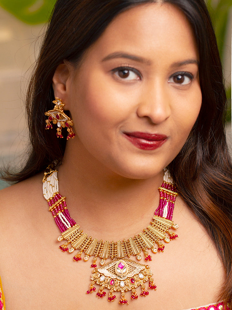 A woman smiles softly wearing the Svarlina Pink Necklace Set, featuring a Kundan necklace with red crystal stones, matching earrings, and a gold-embroidered pink traditional outfit—ideal elegant party wear jewelry.