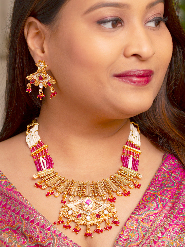 A woman smiles softly wearing the Svarlina Pink Necklace Set, featuring a Kundan necklace with red crystal stones, matching earrings, and a gold-embroidered pink traditional outfit—ideal elegant party wear jewelry.