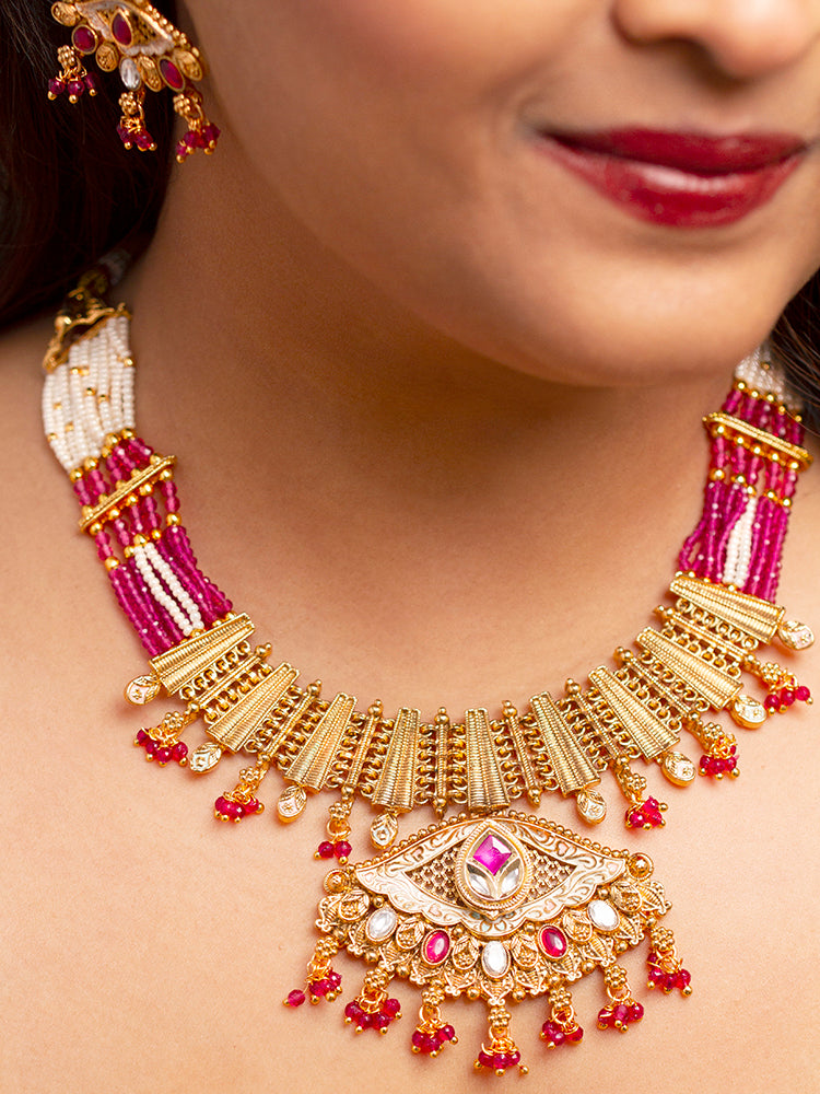 A woman smiles softly wearing the Svarlina Pink Necklace Set, featuring a Kundan necklace with red crystal stones, matching earrings, and a gold-embroidered pink traditional outfit—ideal elegant party wear jewelry.