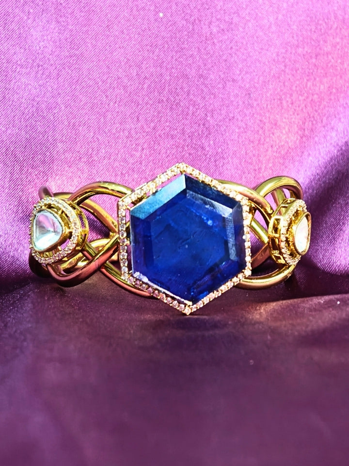 The Tahira Navy Blue Bracelet (Adjustable) features a large hexagonal Navy Blue stone at the center, with an american diamond border and two round kundan stones on each side, displayed on purple satin fabric with white flowers in the background.