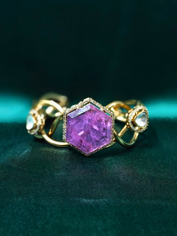 The Tahira Purple Bracelet (Adjustable) features a large hexagonal Purple stone at the center, with an american diamond border and two round kundan stones on each side, displayed on Green satin fabric with white flowers in the background.