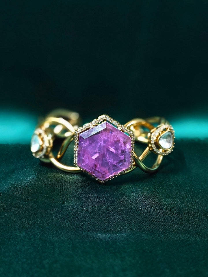 The Tahira Purple Bracelet (Adjustable) features a large hexagonal Purple stone at the center, with an american diamond border and two round kundan stones on each side, displayed on Green satin fabric with white flowers in the background.