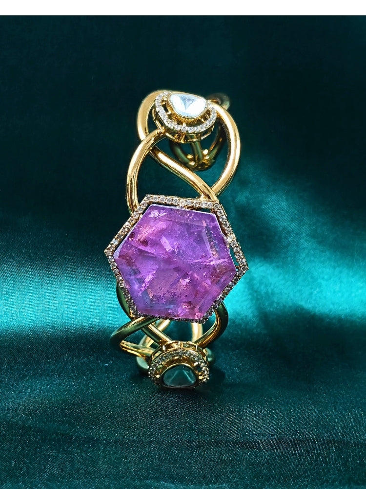 The Tahira Purple Bracelet (Adjustable) features a large hexagonal Purple stone at the center, with an american diamond border and two round kundan stones on each side, displayed on Green satin fabric with white flowers in the background.