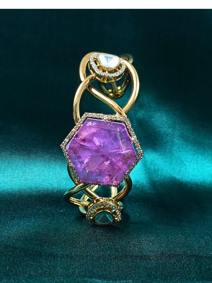 The Tahira Purple Bracelet (Adjustable) features a large hexagonal Purple stone at the center, with an american diamond border and two round kundan stones on each side, displayed on Green satin fabric with white flowers in the background.
