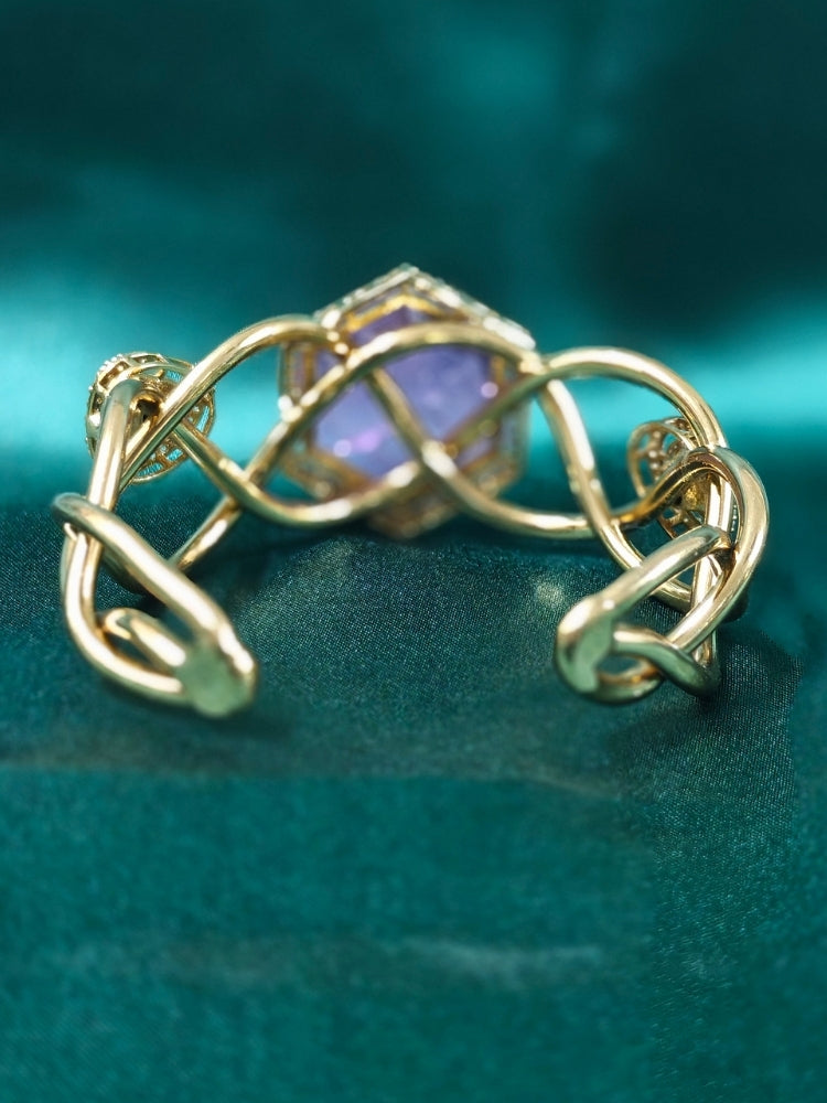 The Tahira Purple Bracelet (Adjustable) features a large hexagonal Purple stone at the center, with an american diamond border and two round kundan stones on each side, displayed on Green satin fabric with white flowers in the background.