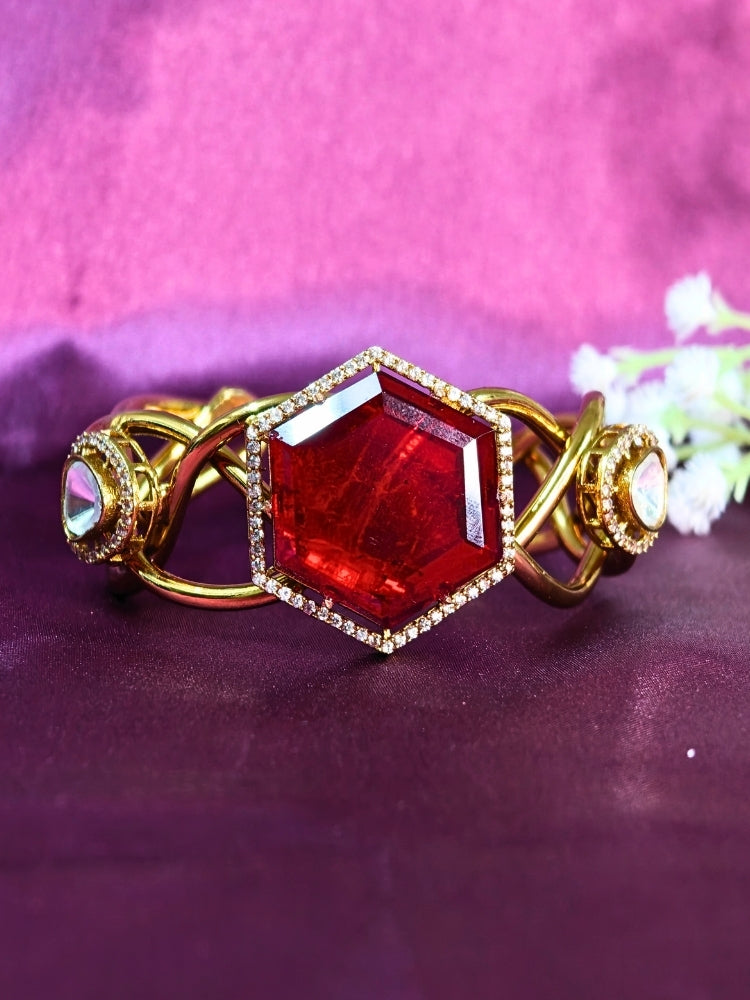 The Tahira Red Bracelet (Adjustable) features a large hexagonal Red stone at the center, with an american diamond border and two round kundan stones on each side, displayed on Purple satin fabric with white flowers in the background.